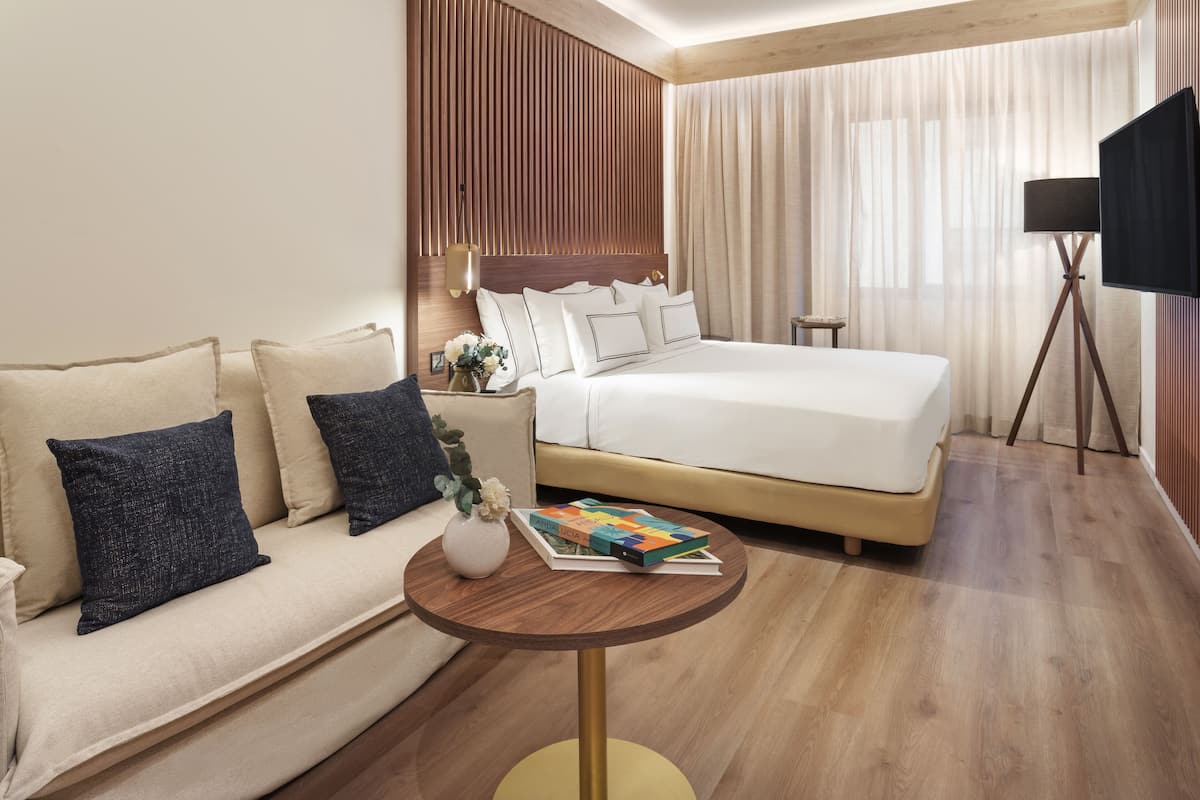 Una habitación de hotel con cama, sofá, una mesita con un libro y televisor sobre un mueble.