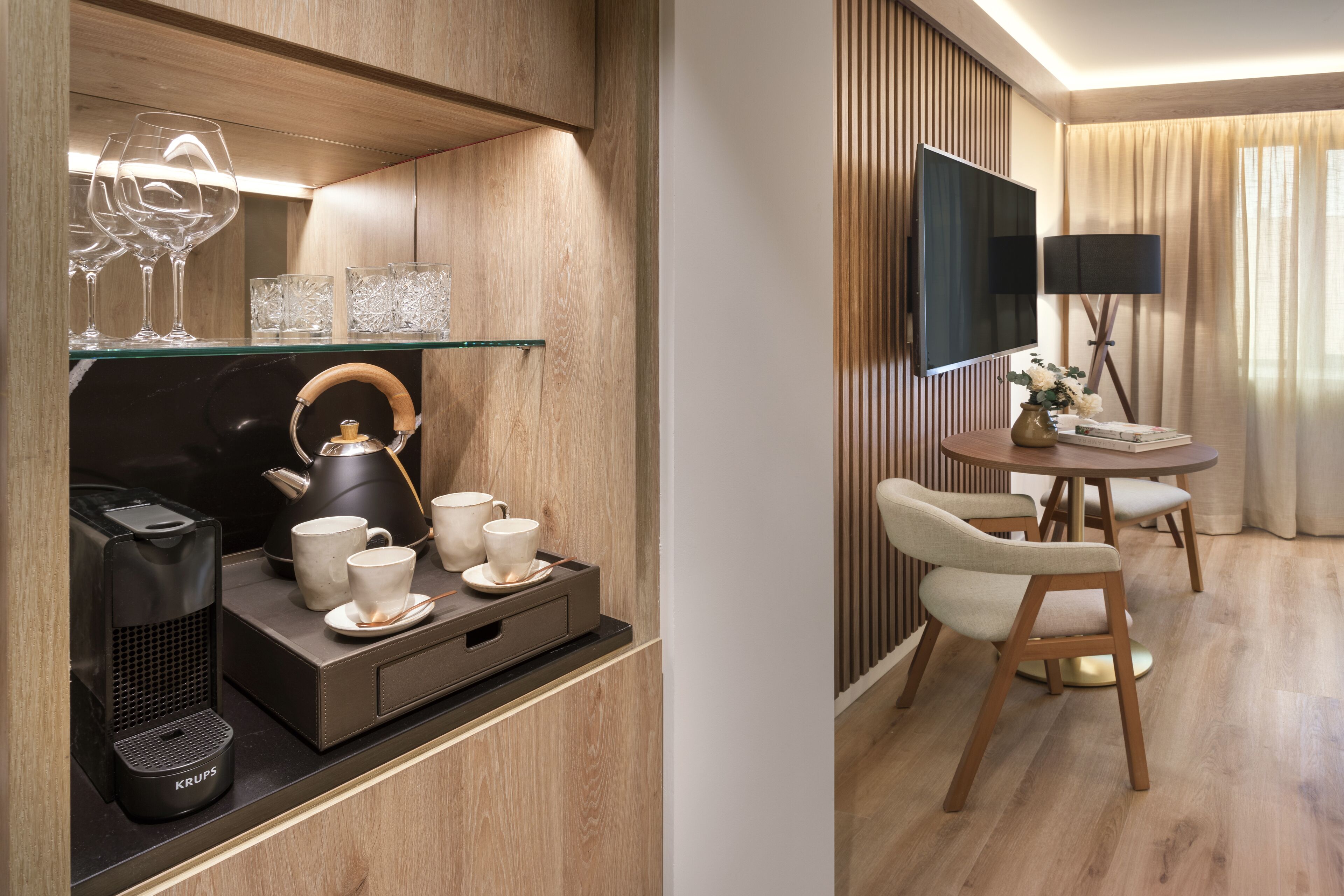 A modern hotel room with a wooden panel wall, a glass shelf with wine glasses and cups, a Krups coffee maker, a round table with chairs, and a TV mounted on the wall.