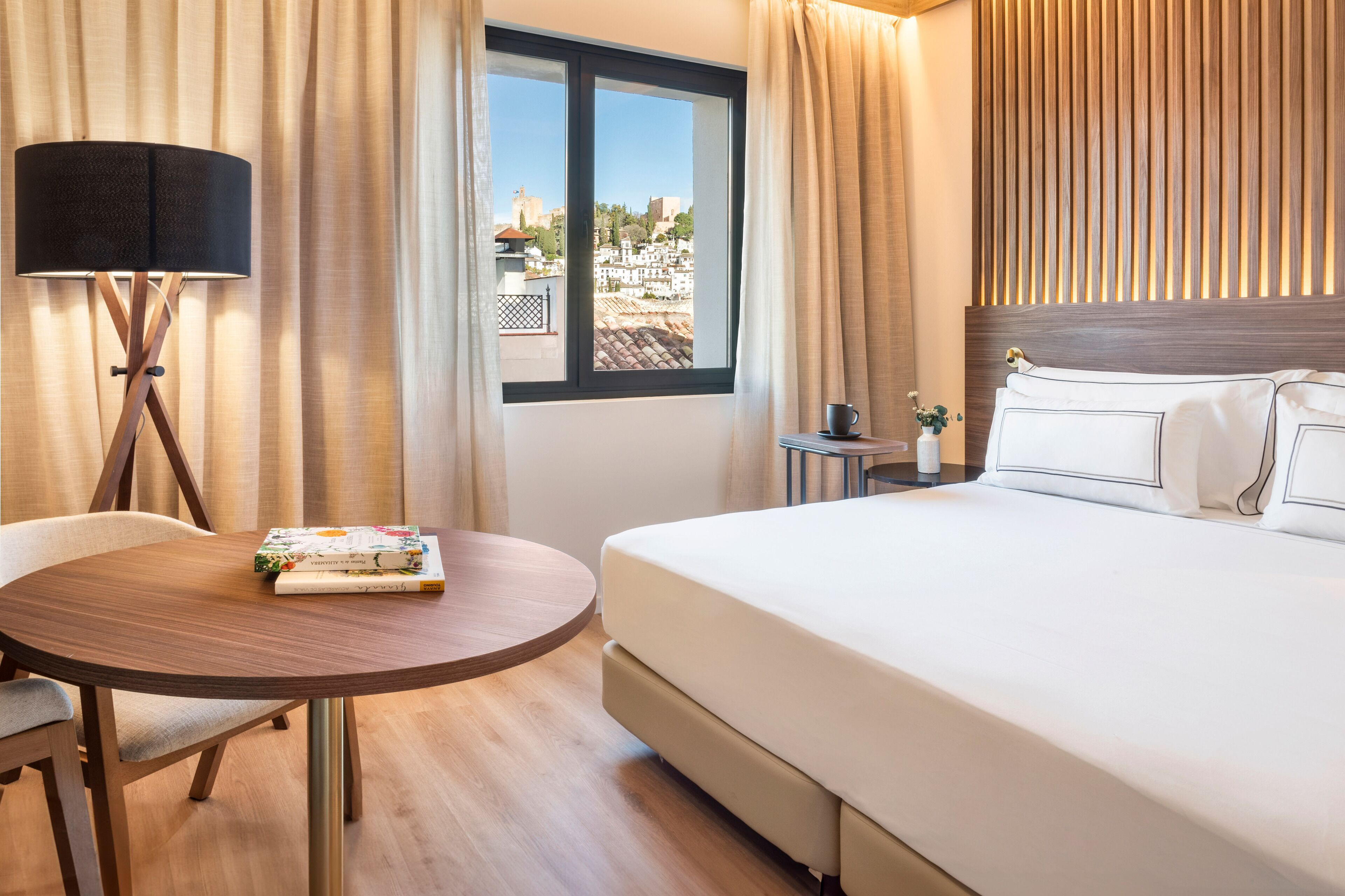 A hotel room with a bed, a round wooden table with a magazine, a lamp, and a window with a view of buildings.