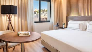 Een hotelkamer met een bed, een ronde houten tafel met een tijdschrift, een lamp en een raam met uitzicht op gebouwen.