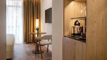 A hotel room with a bed, a small round table with chairs, a TV mounted on the wall, and a wooden shelf with a kettle and glasses.