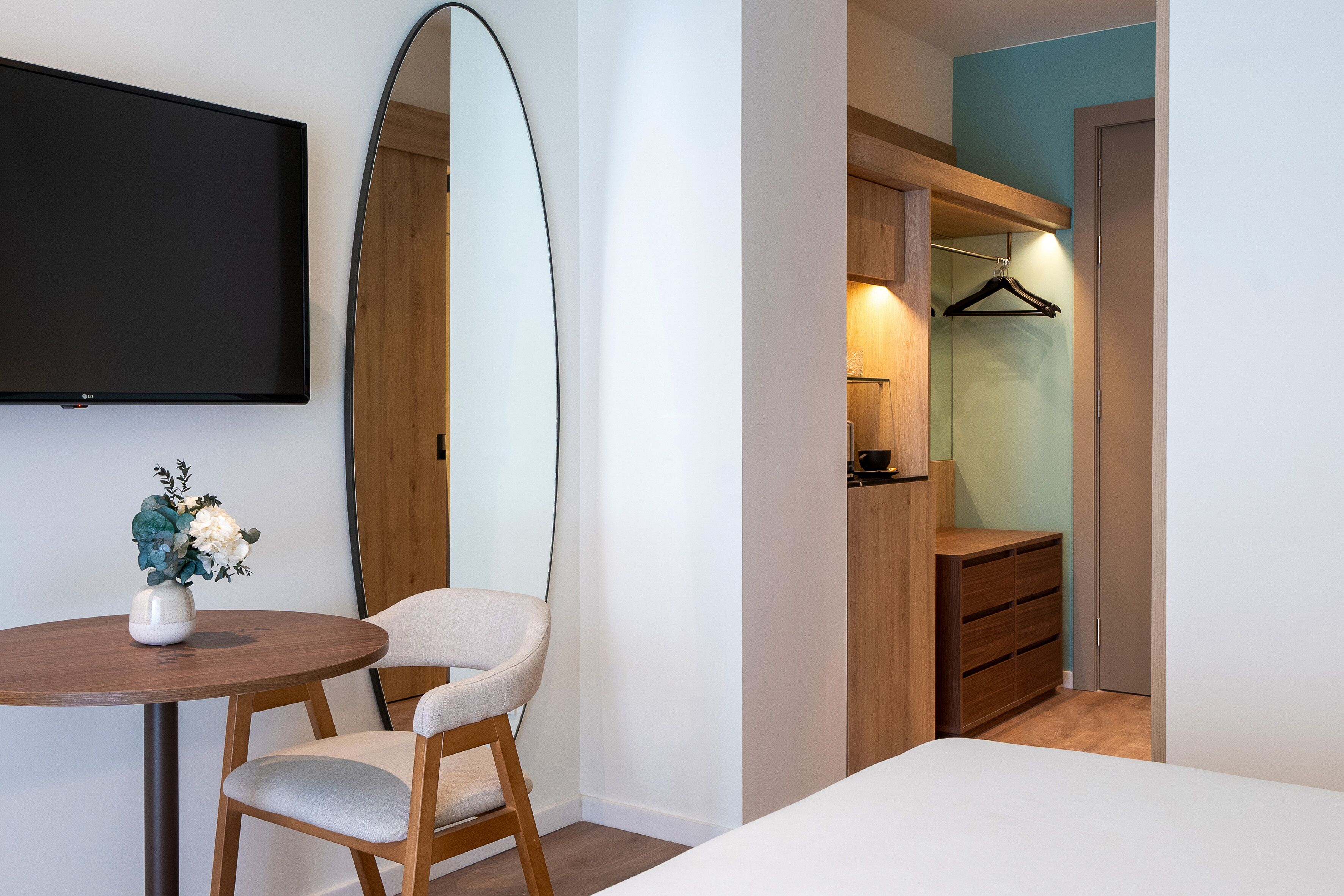 A bedroom with a bed, a chair, a table, a TV, a mirror, and a wardrobe.