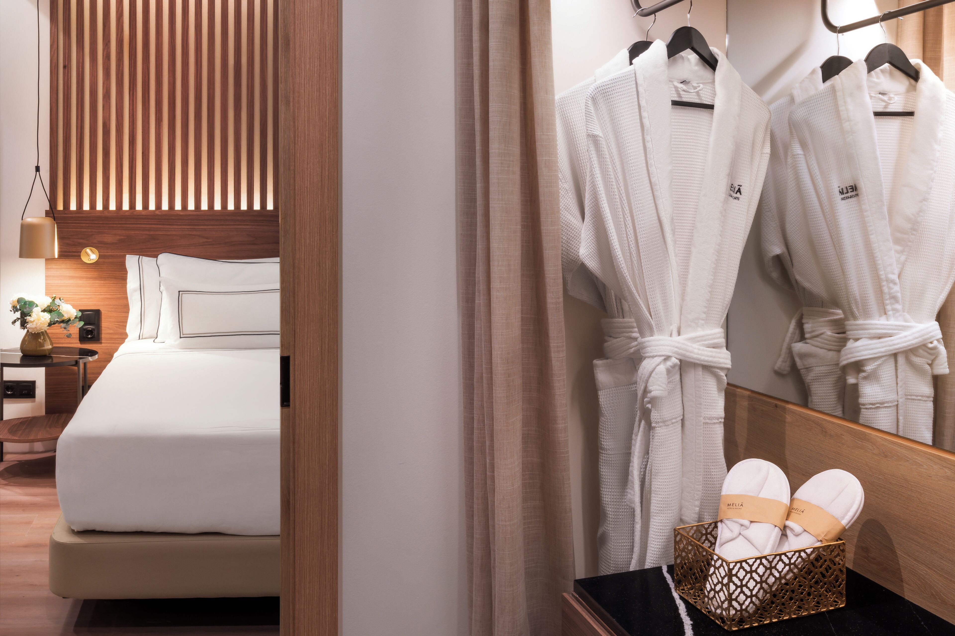 A hotel room with a bed, a nightstand, a lamp, a mirror, and a rack with robes and slippers.