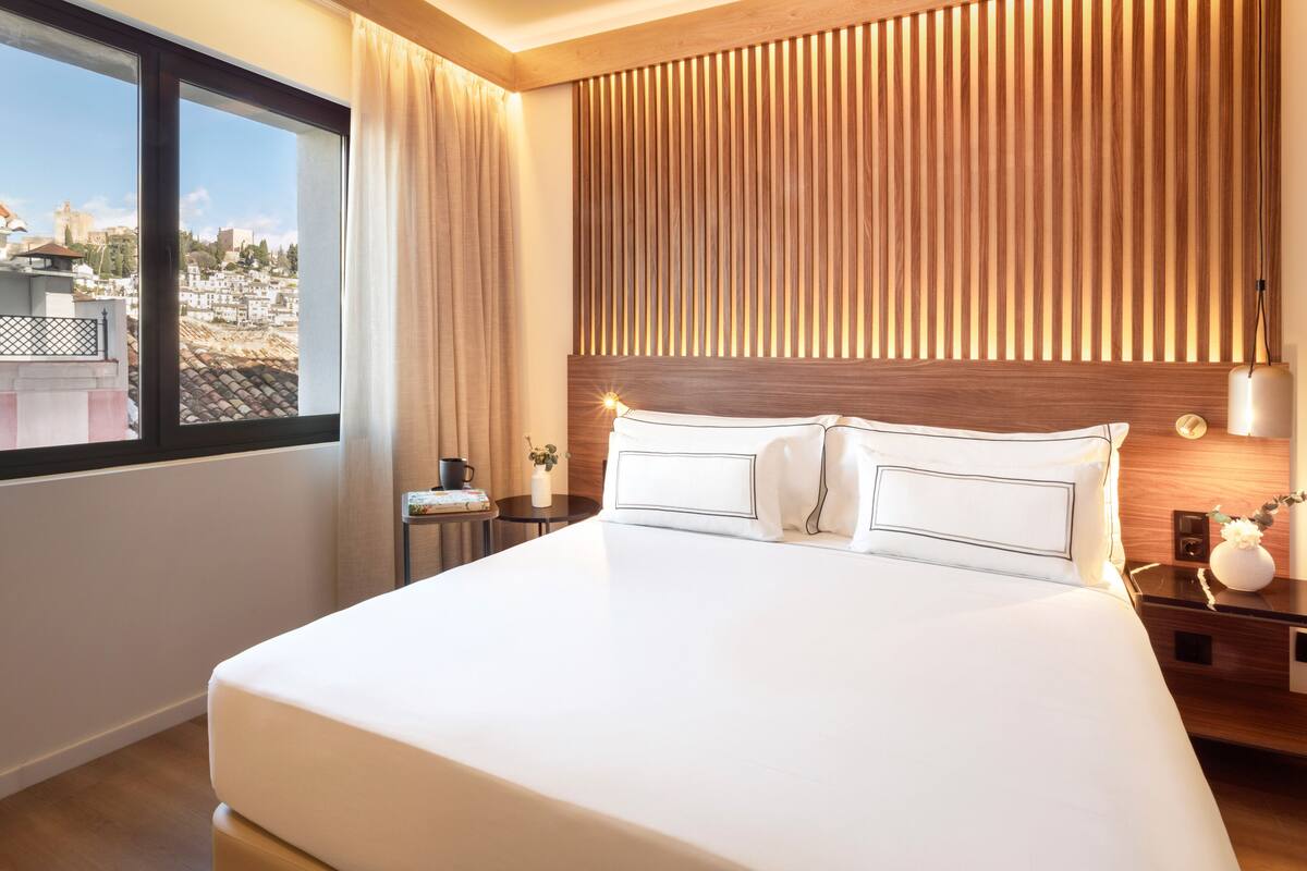 Habitación de hotel con una cama grande, cabecera de madera, mesita de noche con lámpara y vistas a la ciudad a través de una ventana.