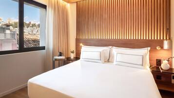 Een hotelkamer met een groot bed, een houten hoofdeinde, een nachtkastje met een lamp en uitzicht op de stad door een raam.