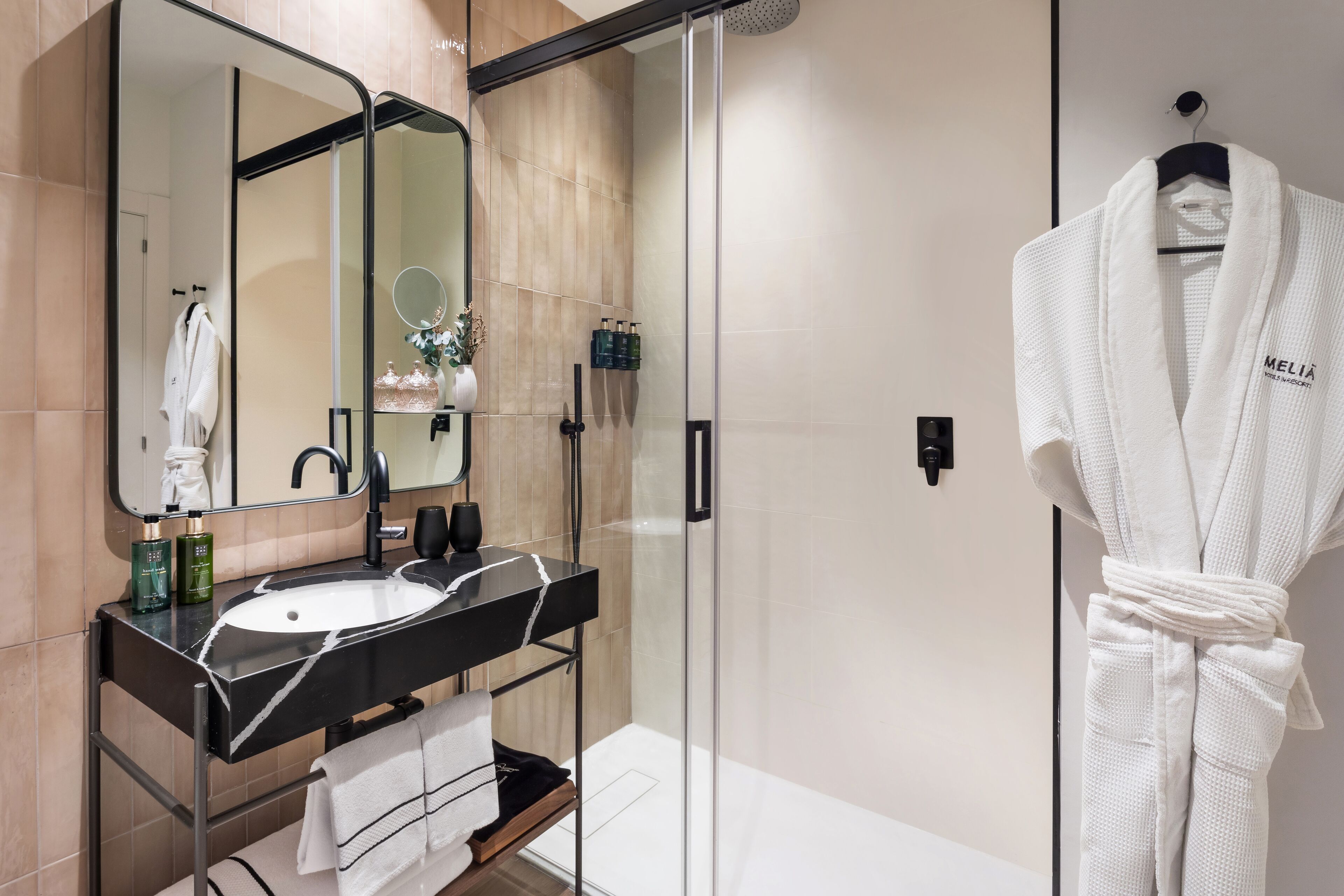 Une salle de bain moderne avec une douche à l’italienne en verre, un meuble-lavabo noir avec un vasque blanche, et un peignoir blanc accroché au mur.