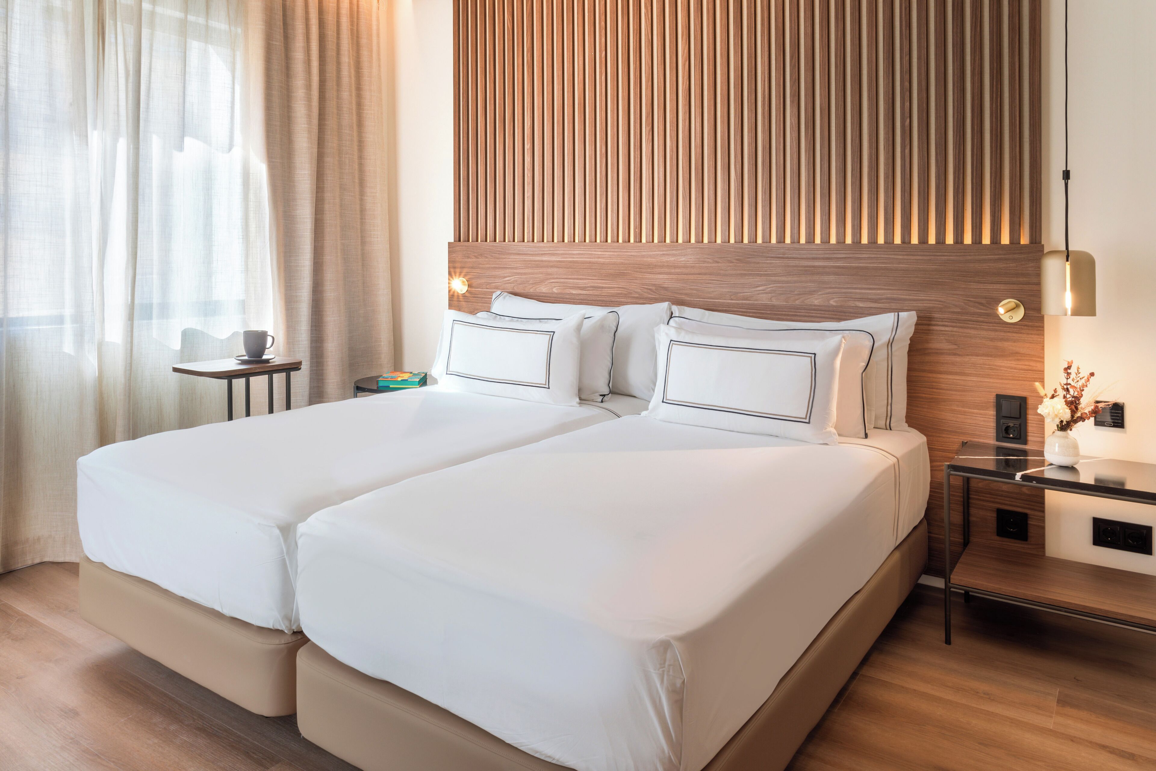 A double bed with white linens, a wooden headboard, a bedside table with a lamp, and a small table with a cup and saucer.