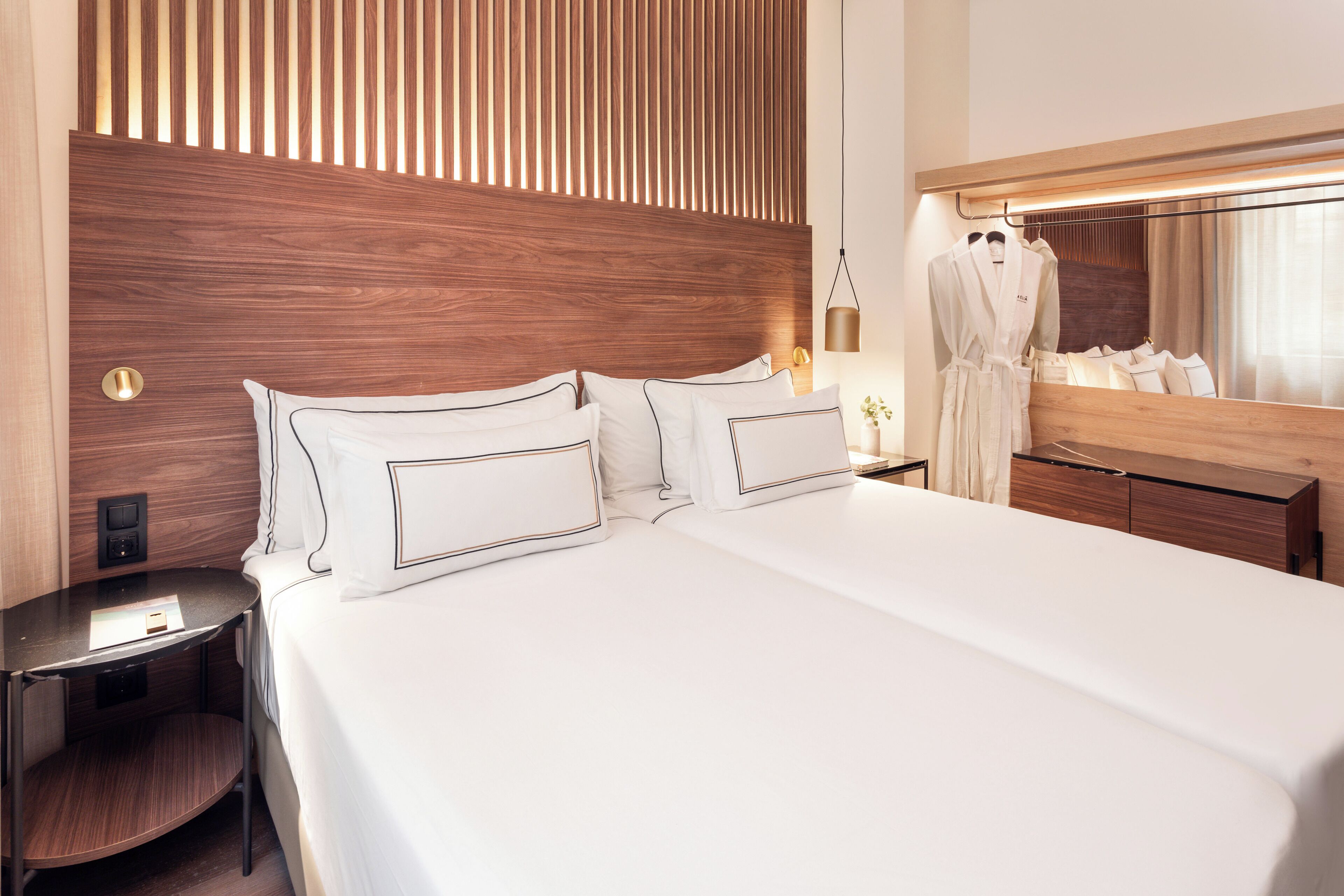 Une chambre d’hôtel moderne avec un grand lit, une tête de lit en bois, une table de chevet, un miroir et un peignoir accroché au mur.