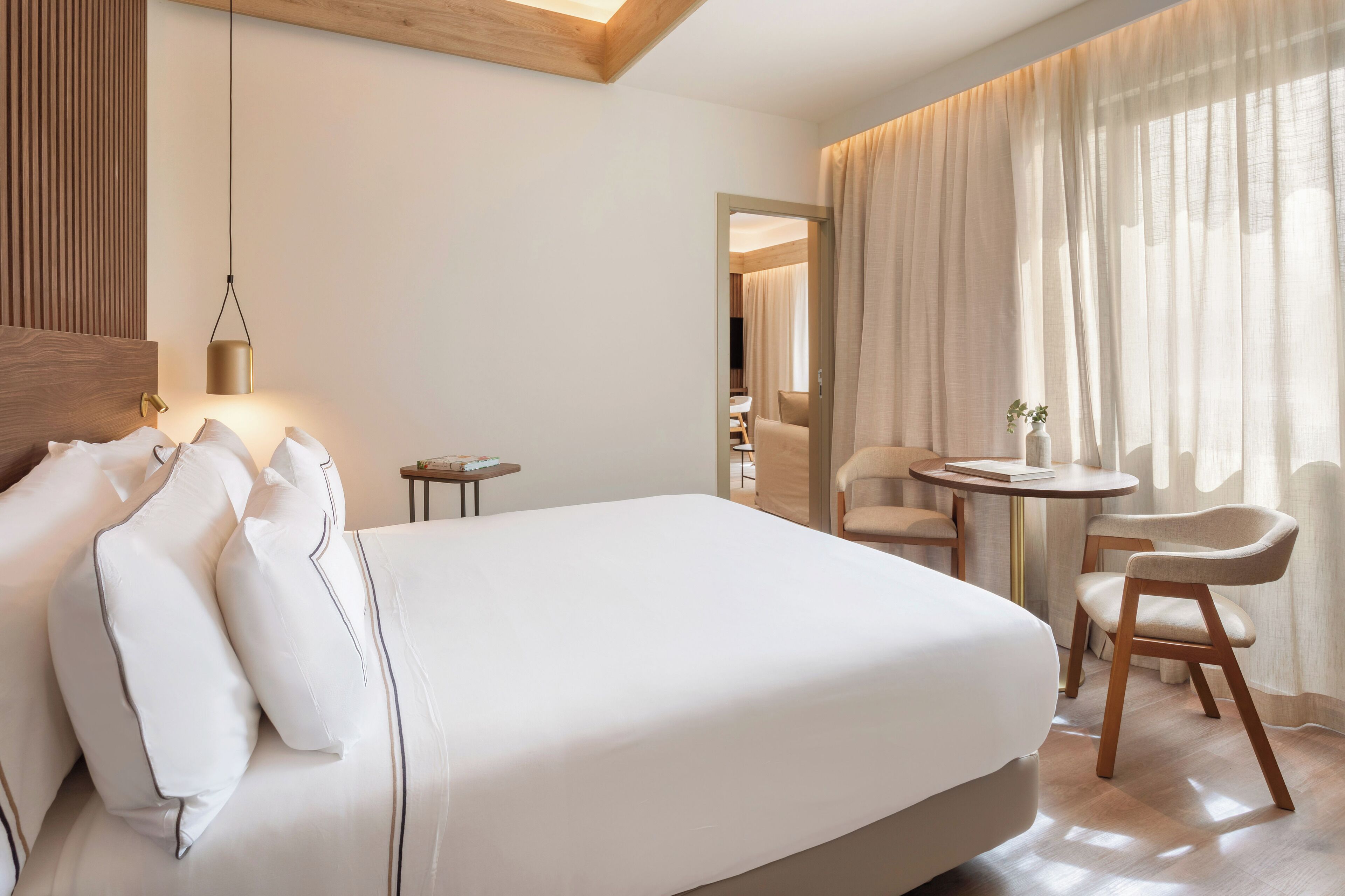 Ett hotellrum med en säng, ett sängbord, en lampa, en stol och utsikt över omgivningen genom gardiner.
