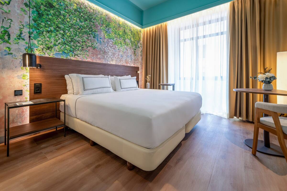 Una habitación de hotel moderna con una cama grande, cabecera de madera, una mesita de noche, una pequeña mesa con un jarrón y vistas al exterior a través de amplias ventanas.