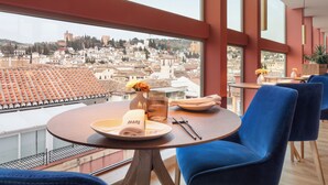 A dining area with a table set for two, a city view, and blue chairs. - Melia Granada Hotel (Granada)