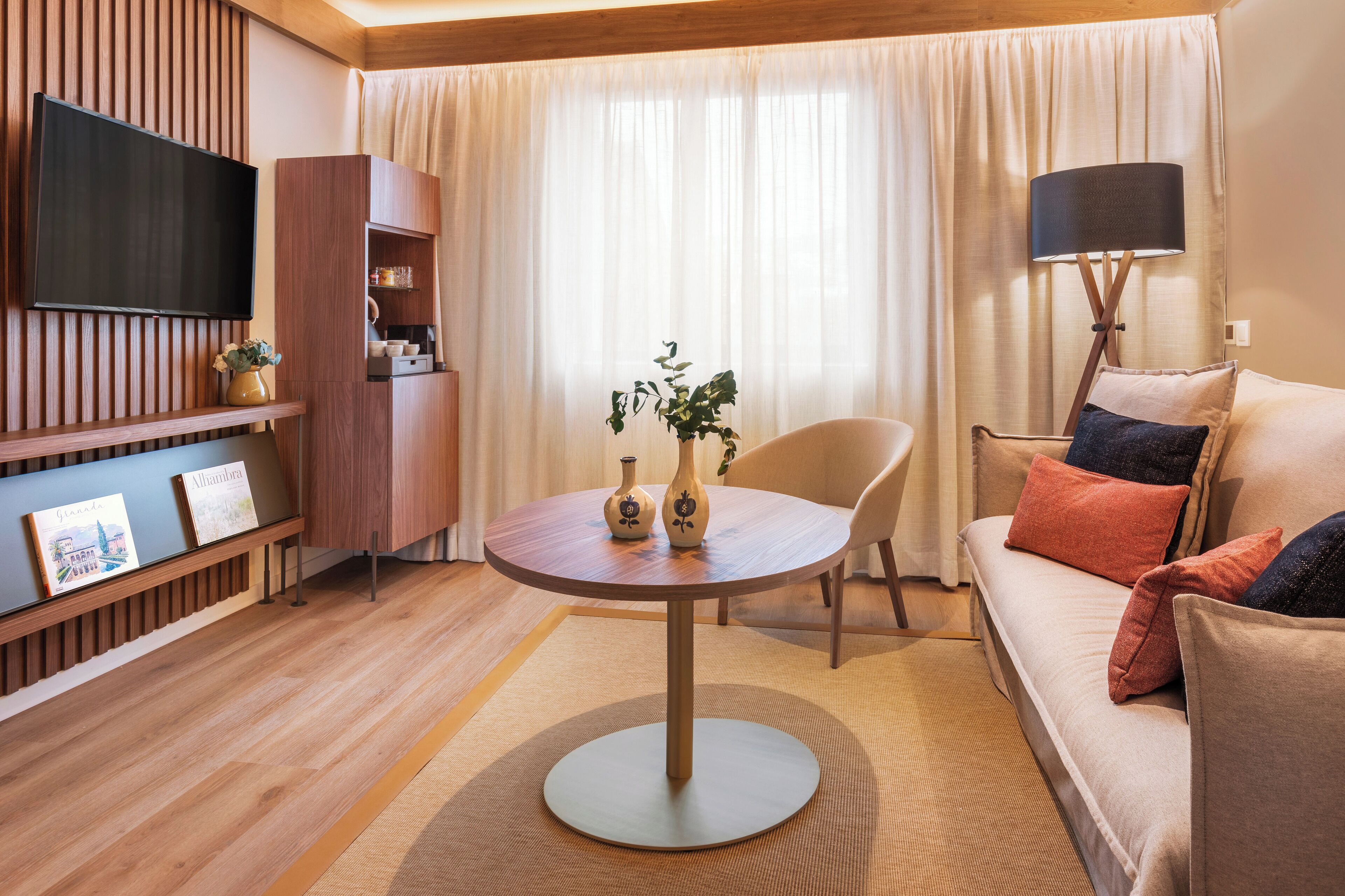 A modern living room with a round table, a sofa, a TV mounted on the wall, and a lamp.