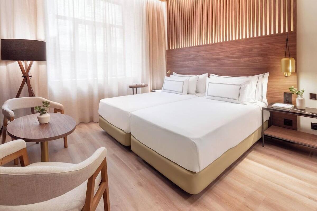 Una cama doble con cabecera de madera, una mesa redonda con sillas, una lámpara y una mesita de noche con una planta.