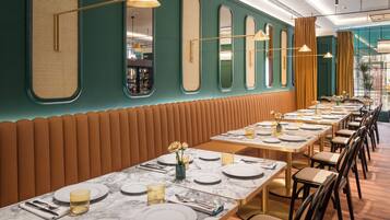 A restaurant with long tables set for dining, featuring a green wall with mirrors and a wooden bench along one side.