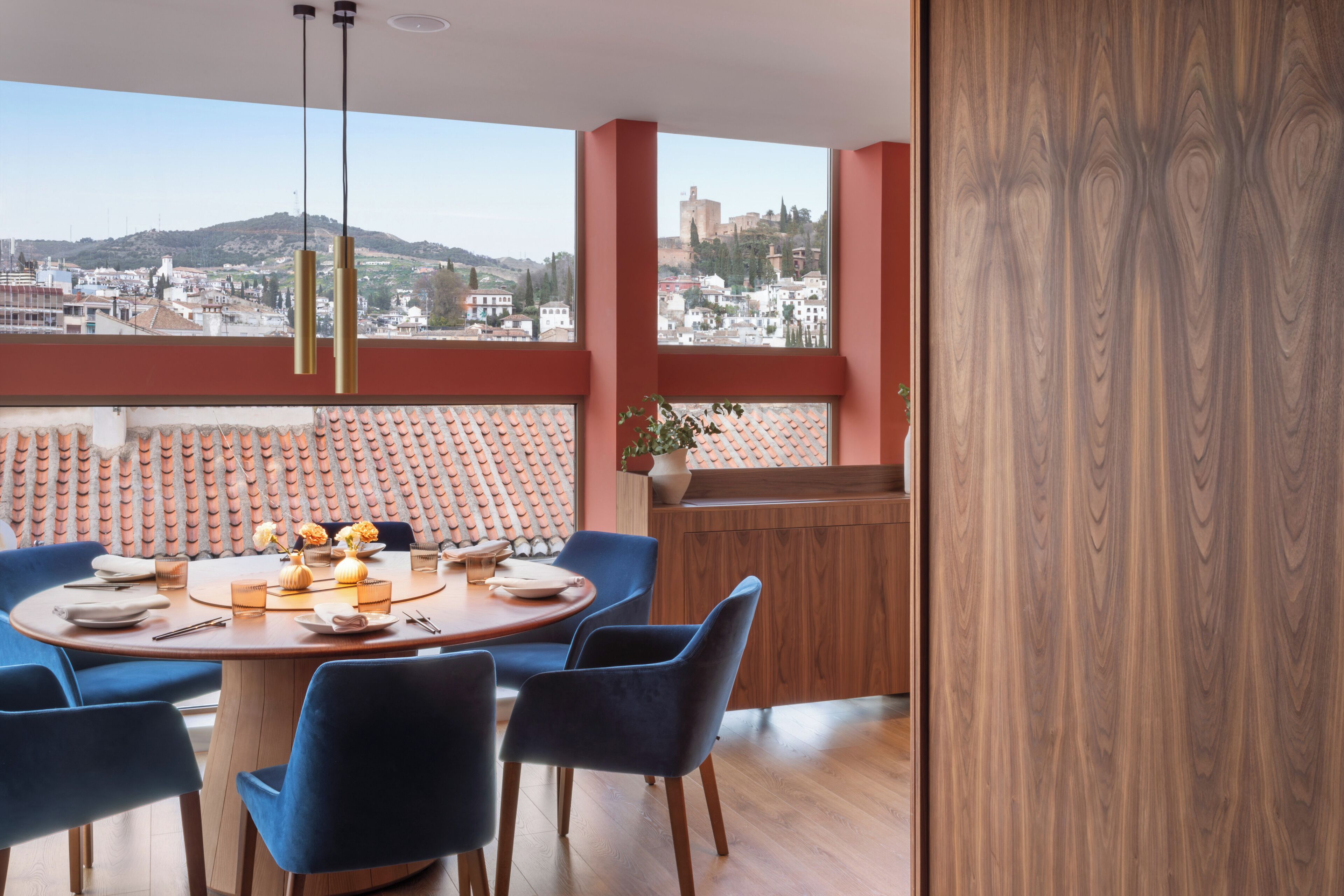 A dining area with a round table, blue chairs, and a view of a cityscape through large windows.
