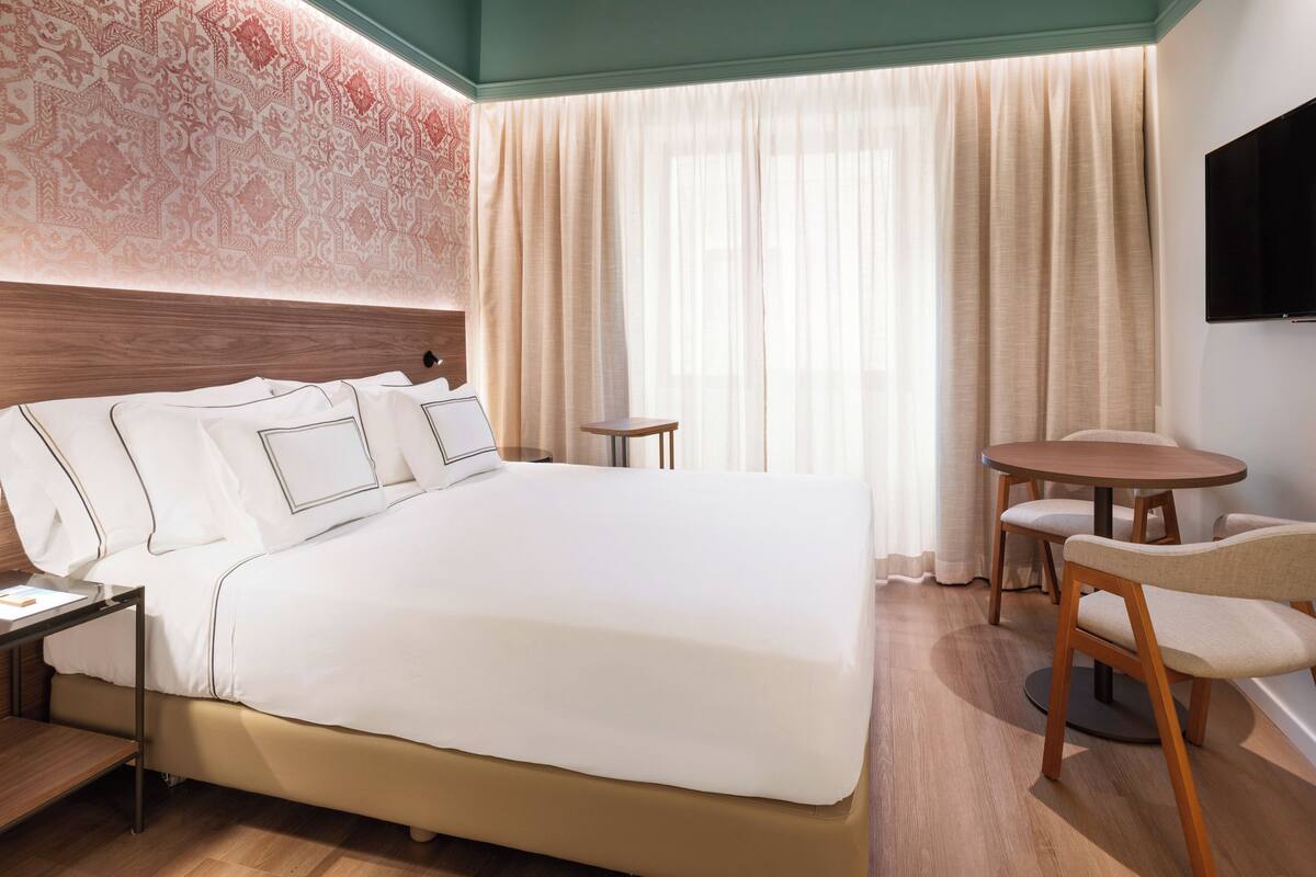 Habitación de hotel con una cama grande, un escritorio, una silla, una mesita y un televisor montado en la pared.