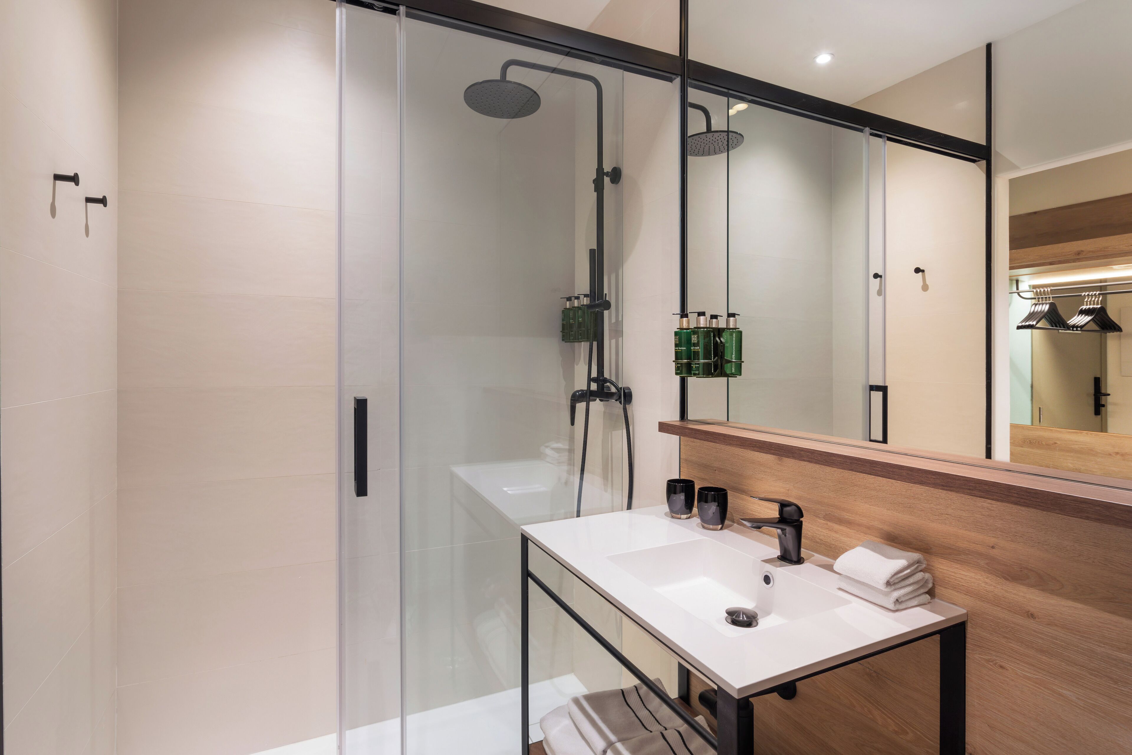 Une salle de bain moderne avec une douche à l’italienne en verre, un lavabo blanc doté d’un plan de toilette en bois et un grand miroir.