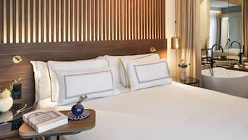 A modern hotel room with a wooden headboard, a large bed with white linens, a bedside table with a book and a decorative vase, and a bathroom visible through a glass partition.