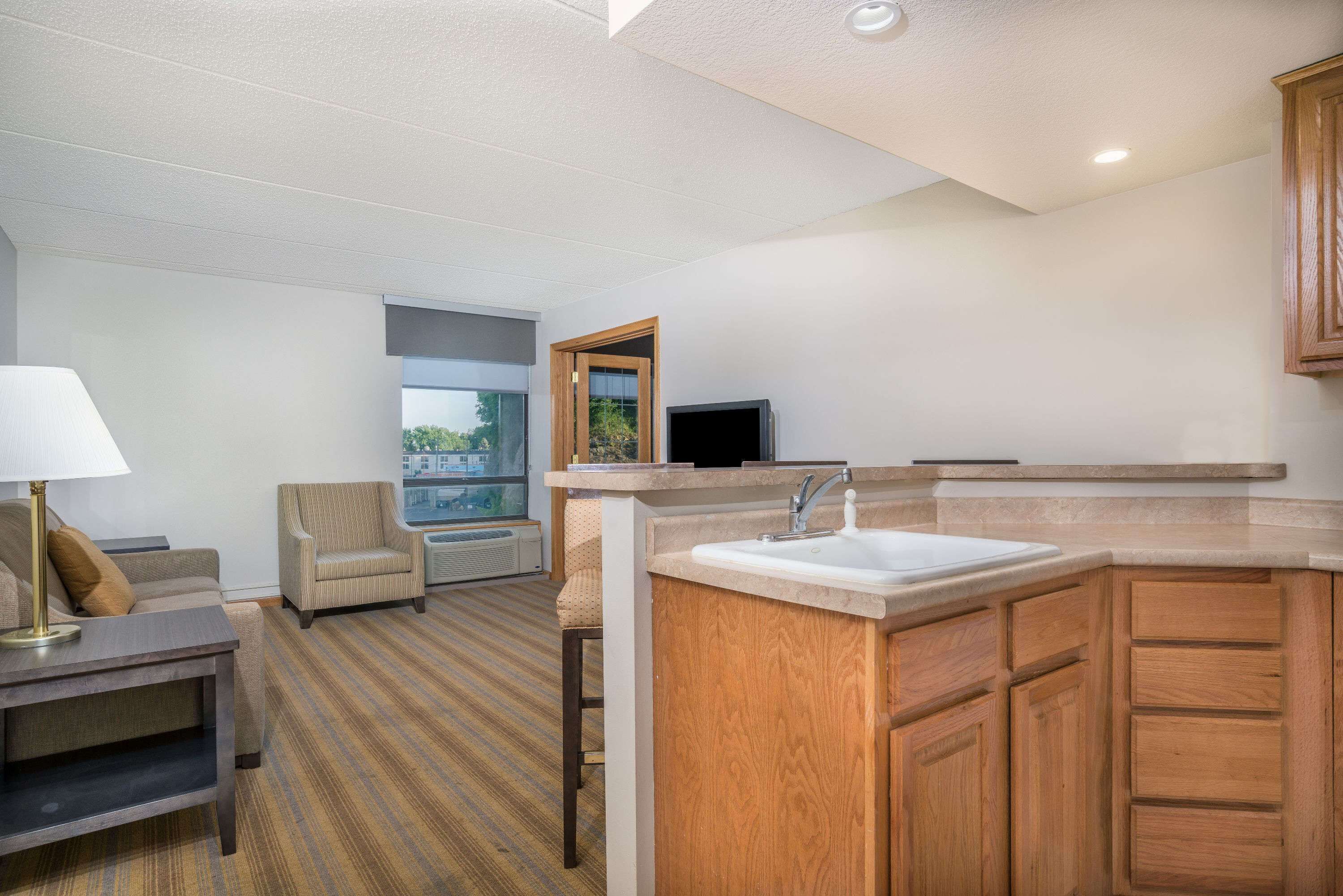A modern hotel room with a sofa, armchair, small table, and a kitchenette area including a sink and wooden cabinets.