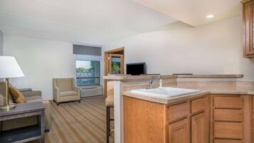 A modern hotel room with a sofa, armchair, small table, and a kitchenette area including a sink and wooden cabinets.