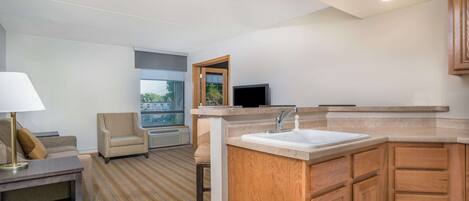 A modern hotel room with a sofa, armchair, small table, and a kitchenette area including a sink and wooden cabinets.