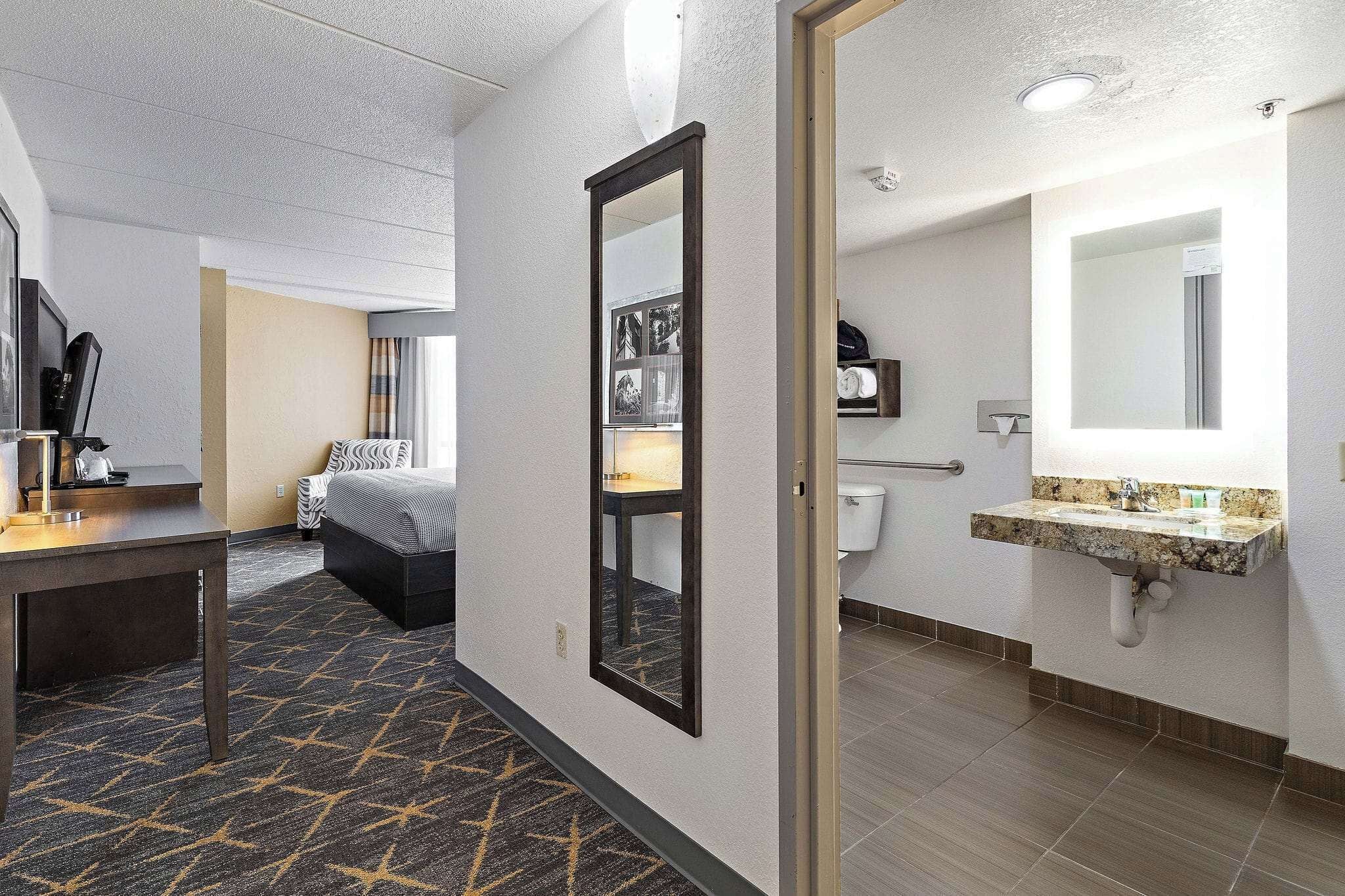 A hotel room with a bed, a desk, a mirror, and a bathroom with a sink and toilet.
