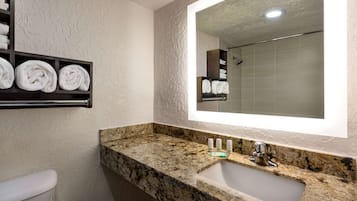 A bathroom with a marble countertop, a sink, and a mirror. There are towels on a shelf above the sink.