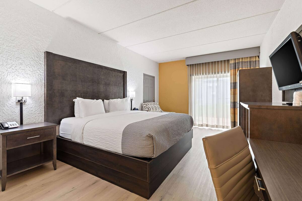 A hotel room with a large bed, a desk with a television, a nightstand, and a window with curtains.