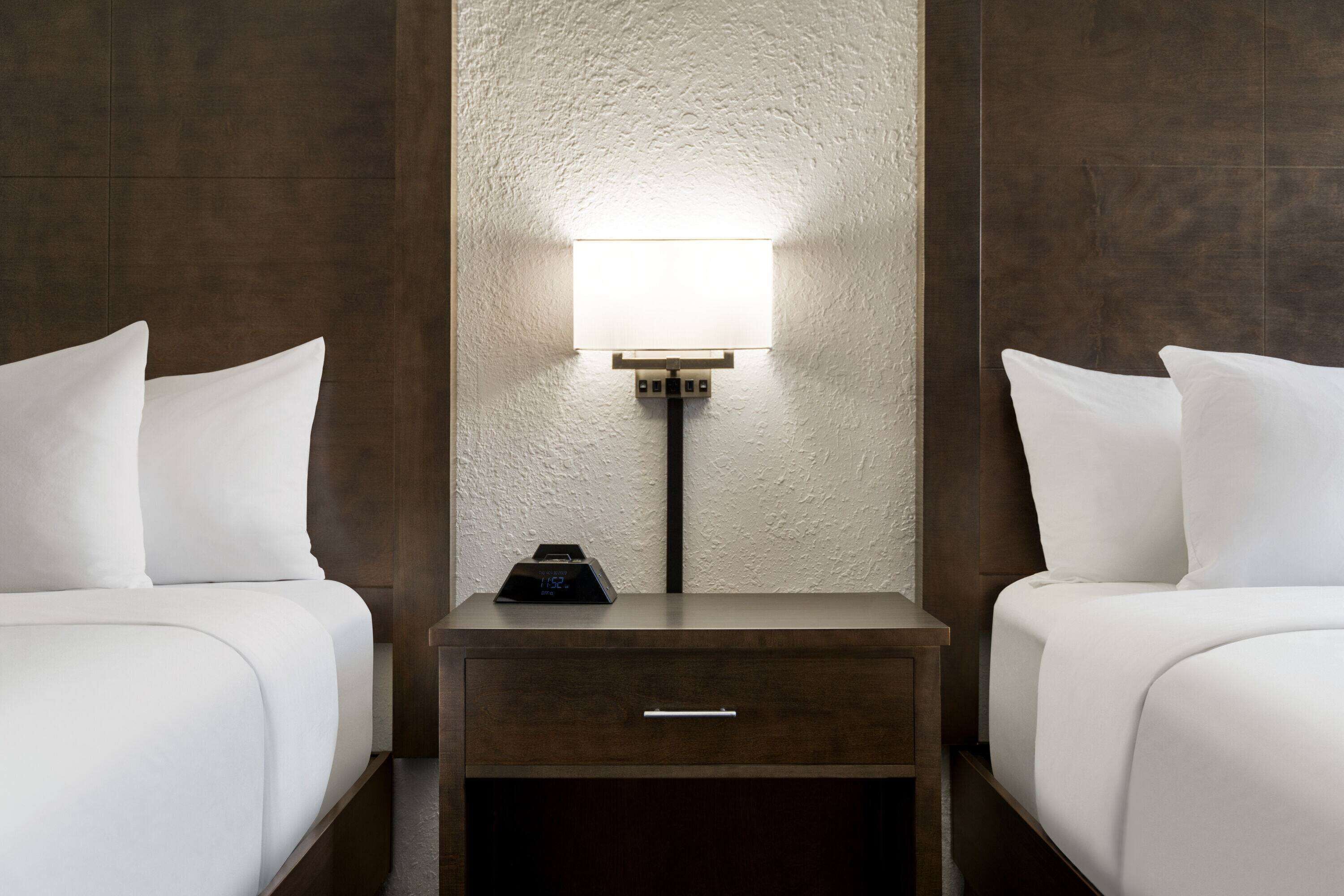 A hotel room with two beds, a nightstand with a lamp, and a digital clock.