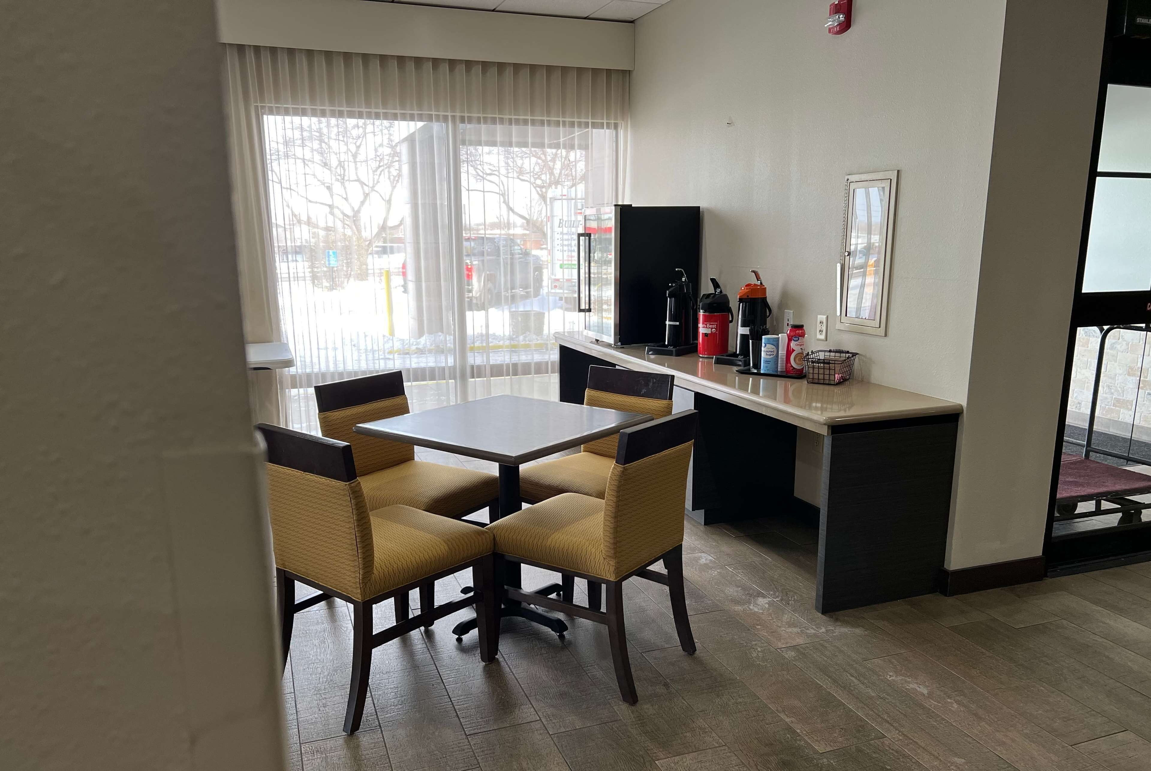 Habitación de hotel con zona de comedor que incluye una mesa y cuatro sillas, una kitchenette pequeña con refrigerador, cafetera y otros electrodomésticos, y un ventanal con persianas.