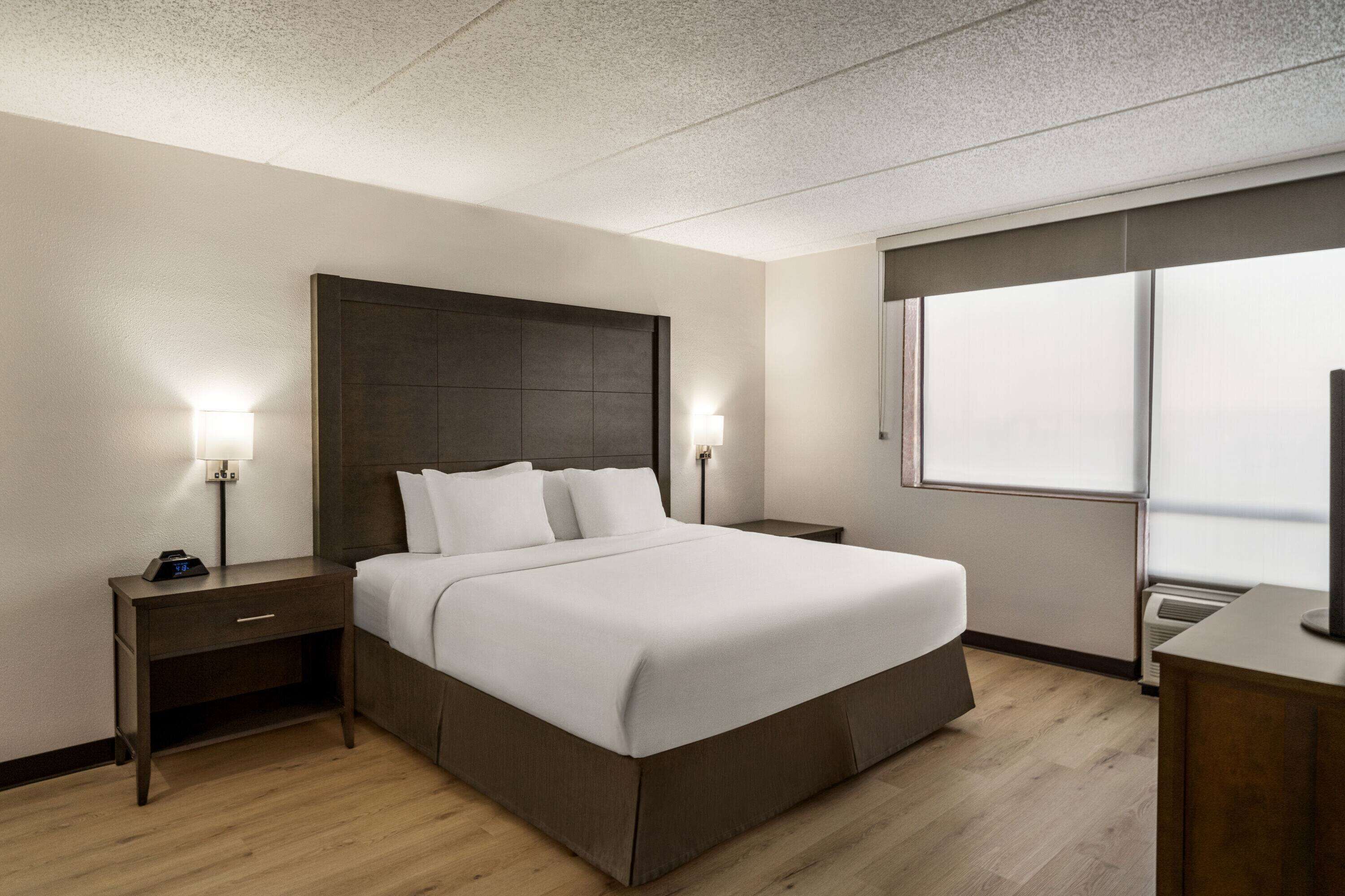 A modern hotel room with a large bed, two bedside tables, a nightstand, a TV, and a window with blinds.