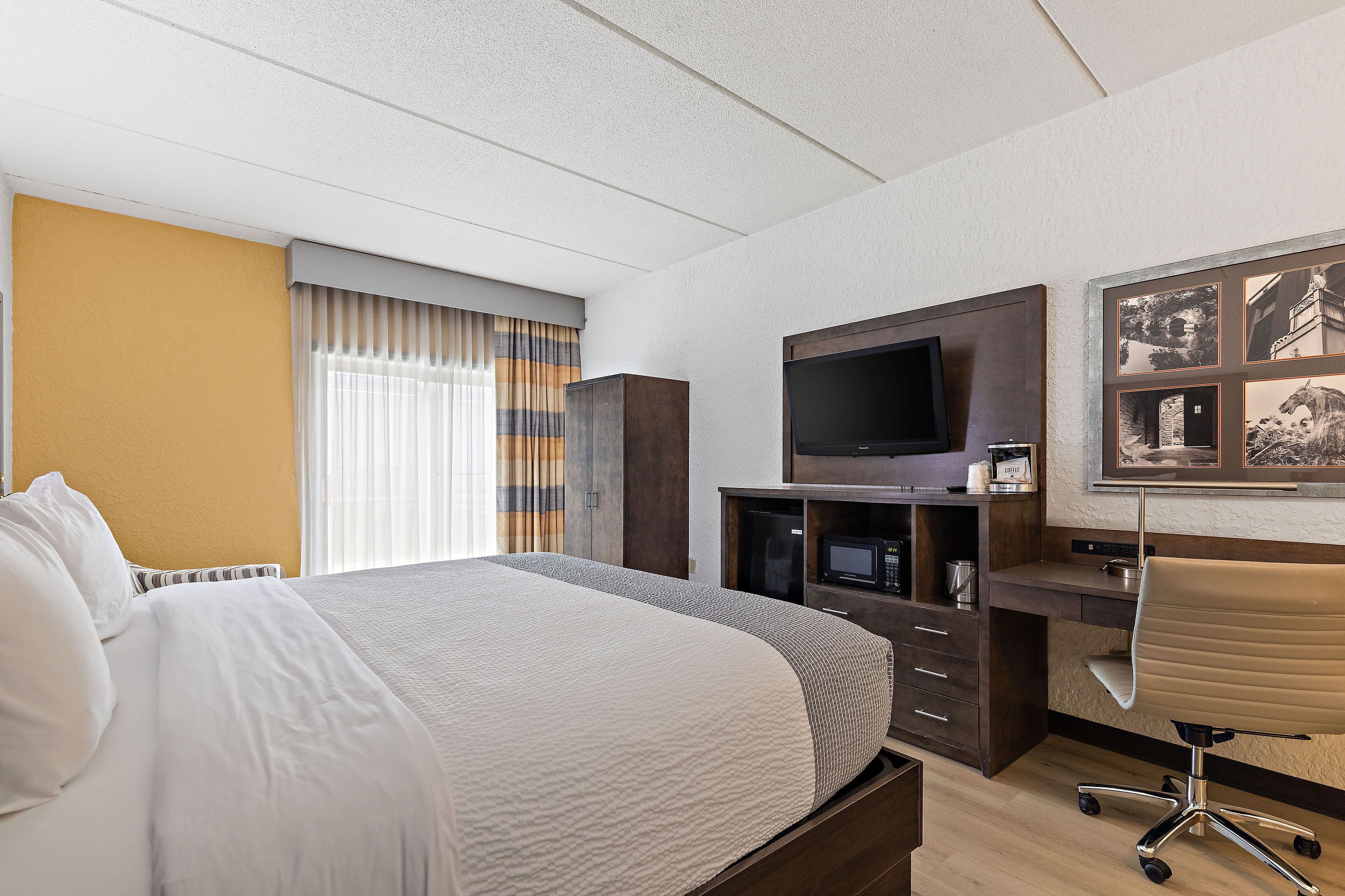 A hotel room with a large bed, a desk with a chair, a flat-screen TV, and a wall-mounted TV.