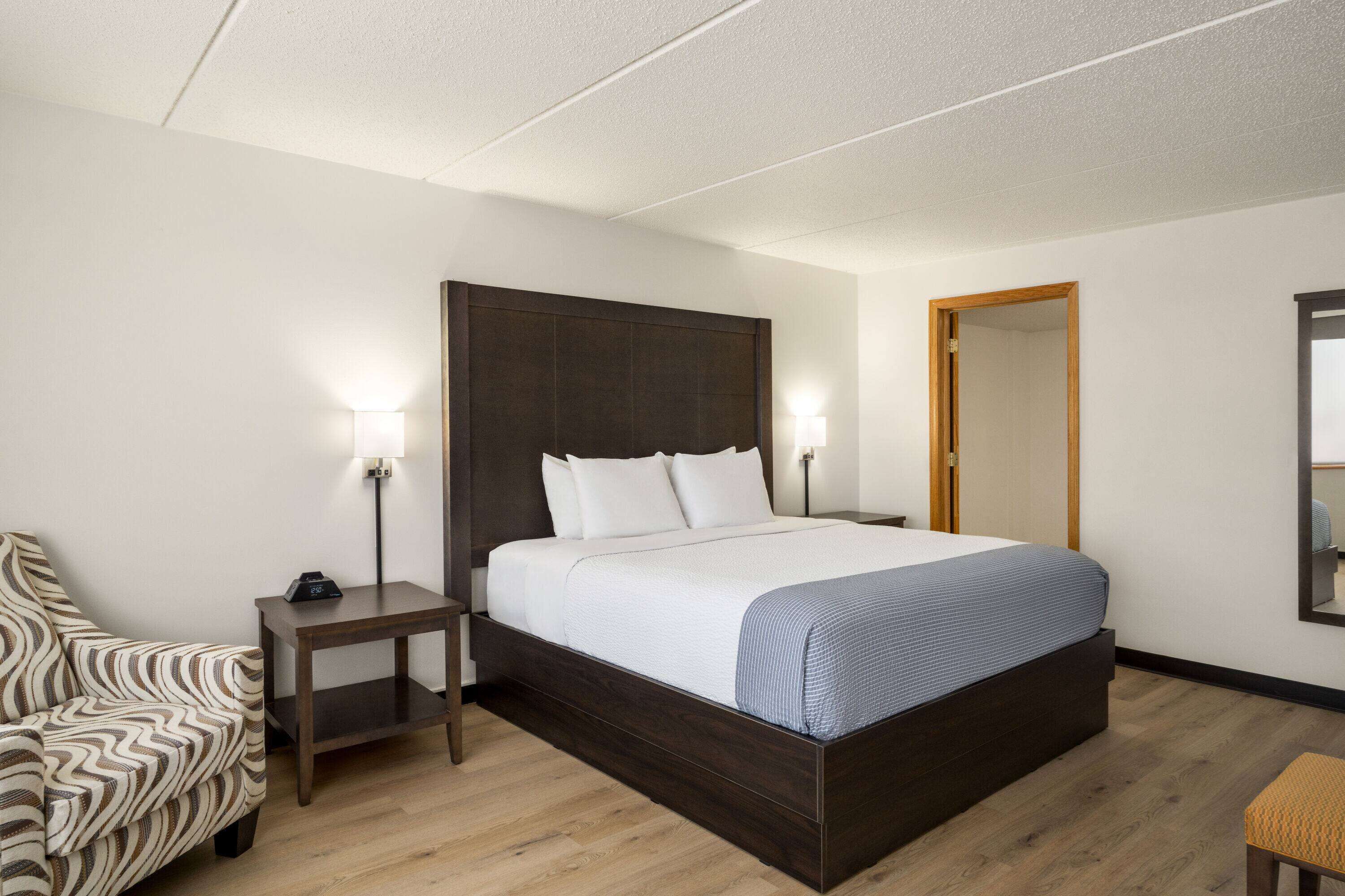 Een hotelkamer met een groot bed, een nachtkastje, een stoel, een kleine tafel en een spiegel aan de muur.