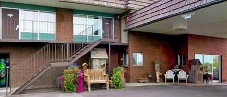 A two-story building with a covered parking area, a staircase leading to the upper floor, and a small outdoor seating area with a bench and potted plants.