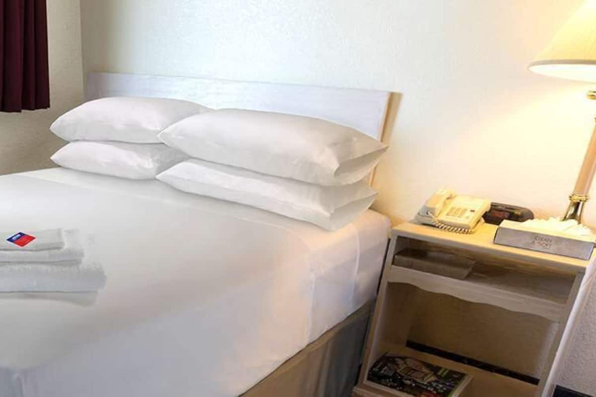 A neatly made bed with white linens and pillows, a bedside table with a lamp and a phone, and a window with curtains.