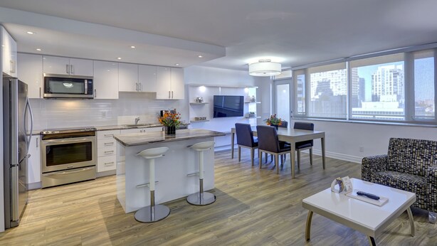 A modern kitchen with white cabinetry, a microwave, and an oven. There is a dining area with a table and chairs, and a patterned sofa. - Town Inn Suites Hotel (Toronto)
