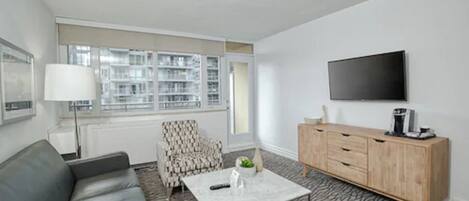 A modern living room with a grey sofa, patterned armchair, coffee table, and a wooden cabinet. A flat-screen TV is mounted on the wall.