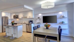 A modern kitchen with white cabinetry, a built-in microwave, and a dishwasher. There is a dining area with a table and chairs, and a TV mounted on the wall. - Town Inn Suites Hotel (Toronto)