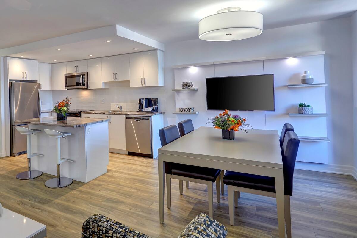 A modern kitchen with white cabinetry, a built-in microwave, and a dishwasher. There is a dining area with a table and chairs, and a TV mounted on the wall.