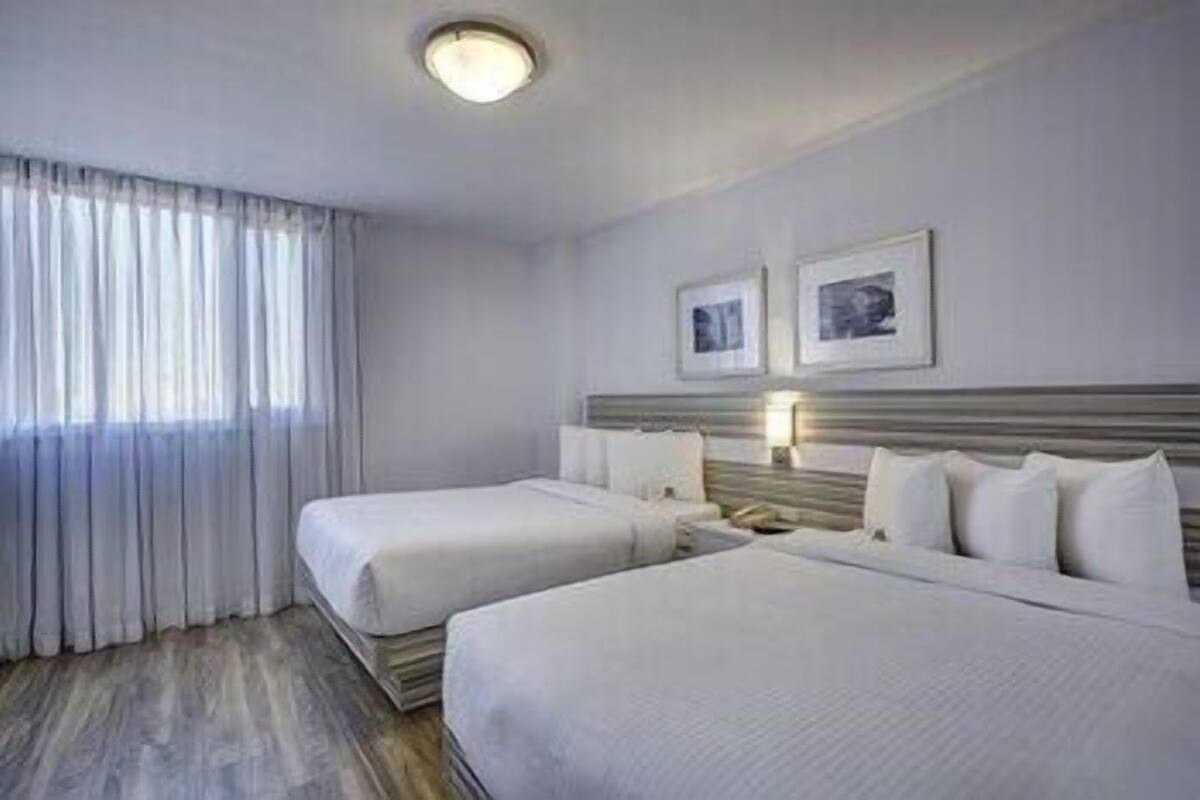 A hotel room with two beds, a nightstand, a lamp, and a window with curtains.