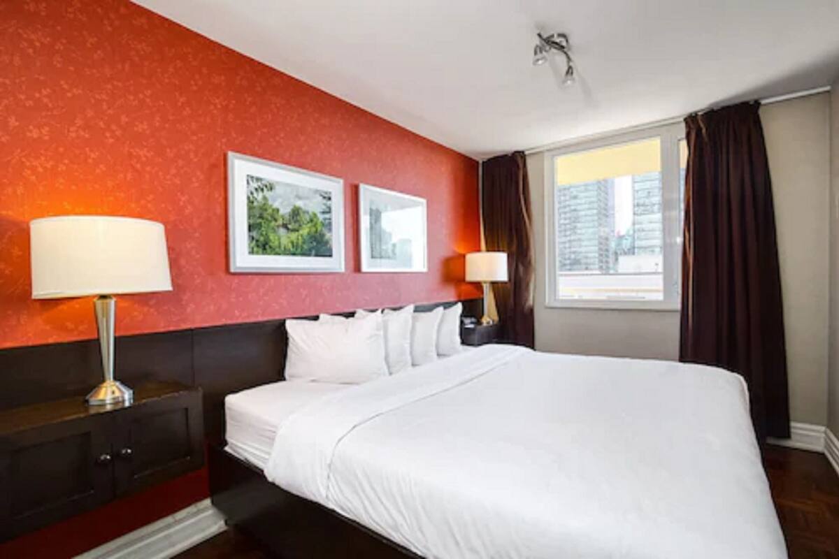 A hotel room with a large bed, two bedside tables with lamps, a red accent wall, and a window with curtains.