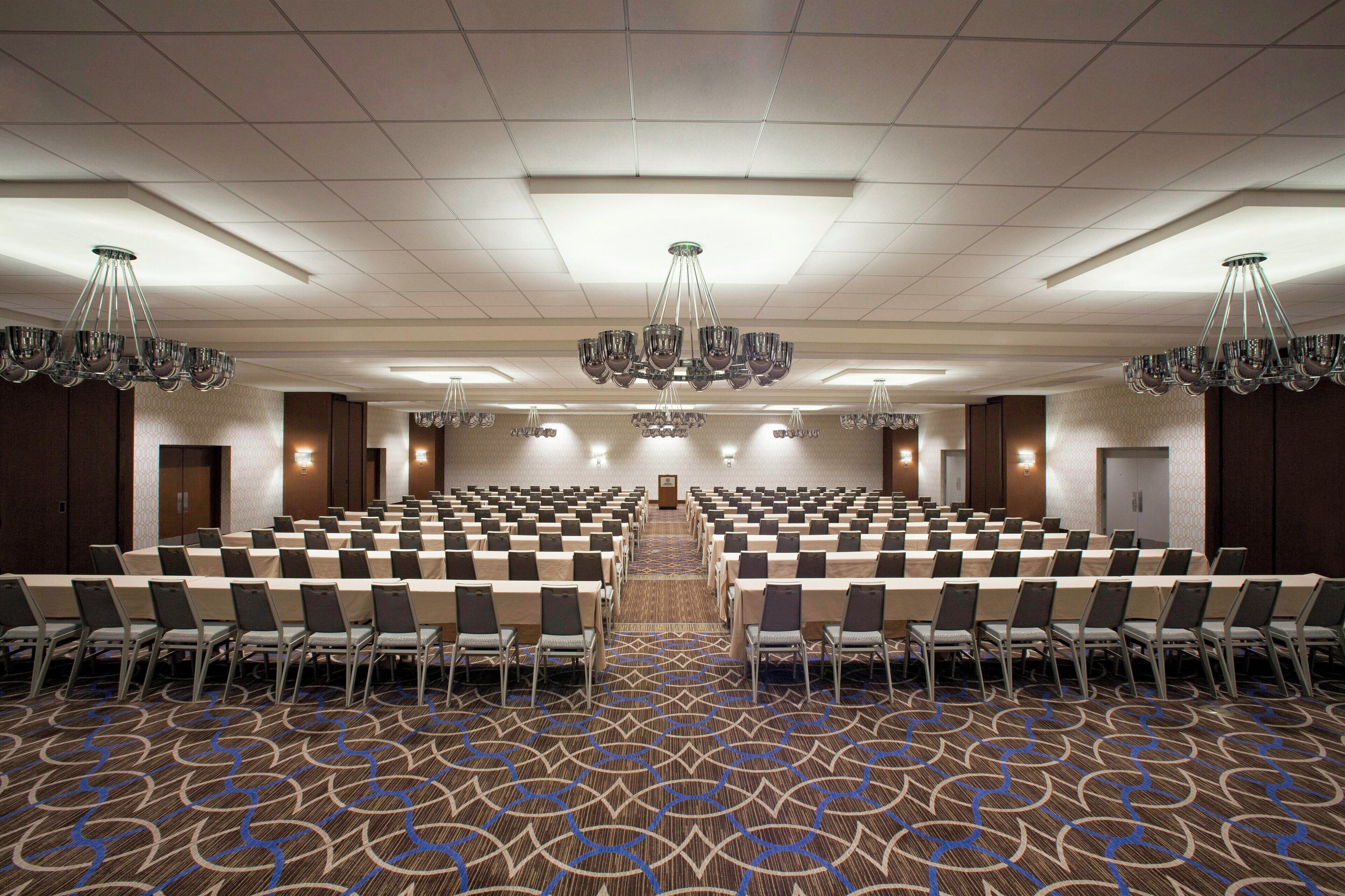 A conference room with rows of tables and chairs, a patterned carpet, and chandeliers.
