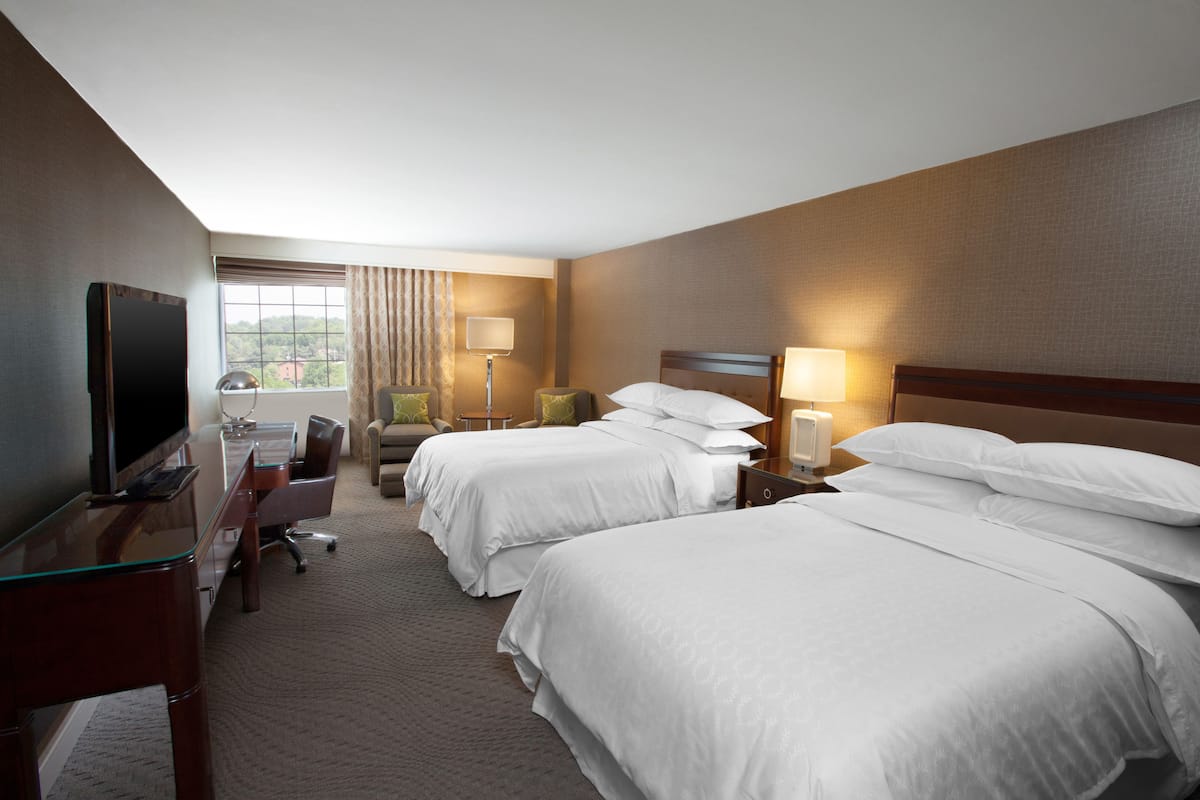 A hotel room with two beds, a desk with a TV, a chair, a lamp, and a window with curtains.