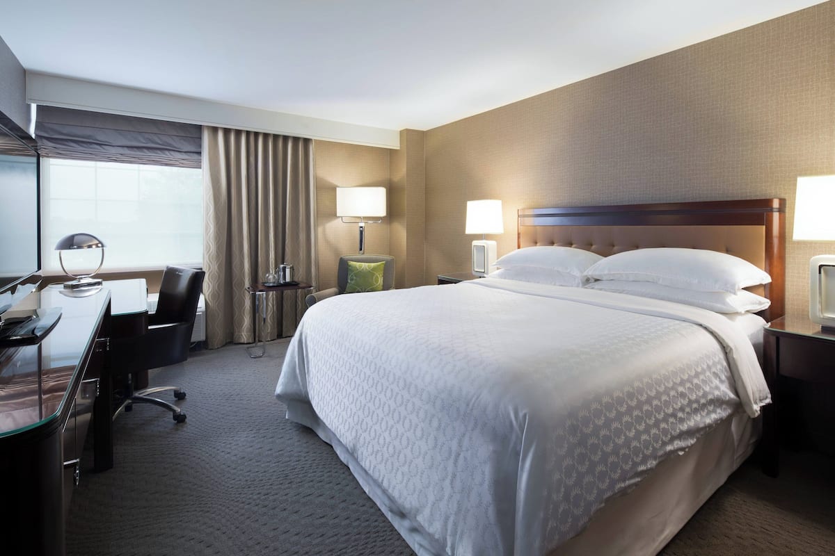 A hotel room with a large bed, a desk with a chair, a lamp, and a window with curtains.