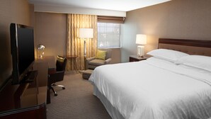 A hotel room with a bed, a desk with a television, a chair, a lamp, and a window with curtains.
