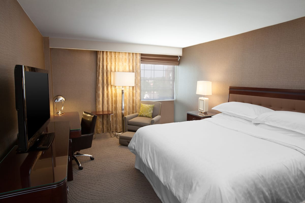 A hotel room with a bed, a desk with a television, a chair, a lamp, and a window with curtains.