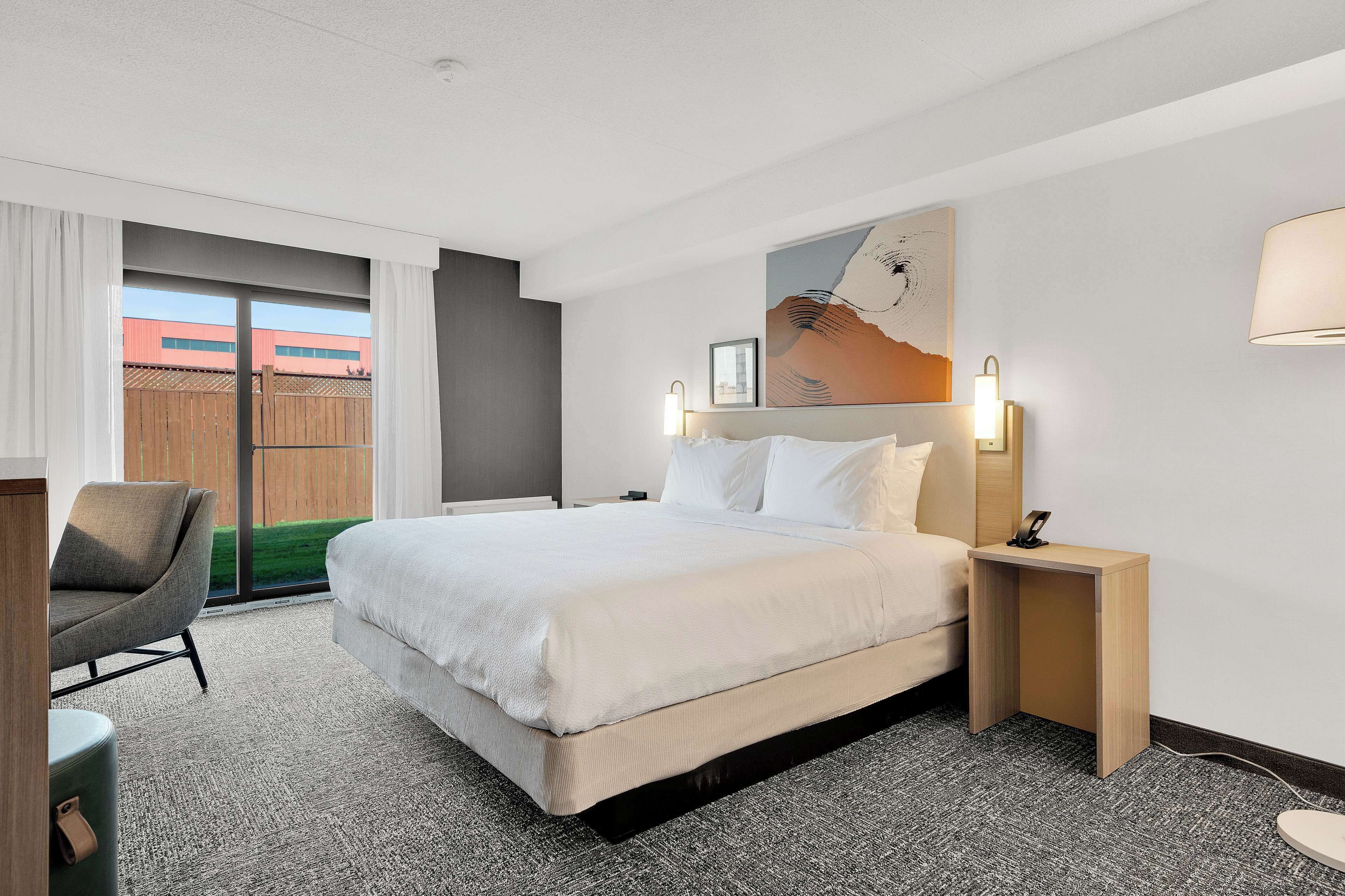 Habitación de hotel con una cama grande, un escritorio, una silla, una mesita de noche con lámpara y vistas al exterior.