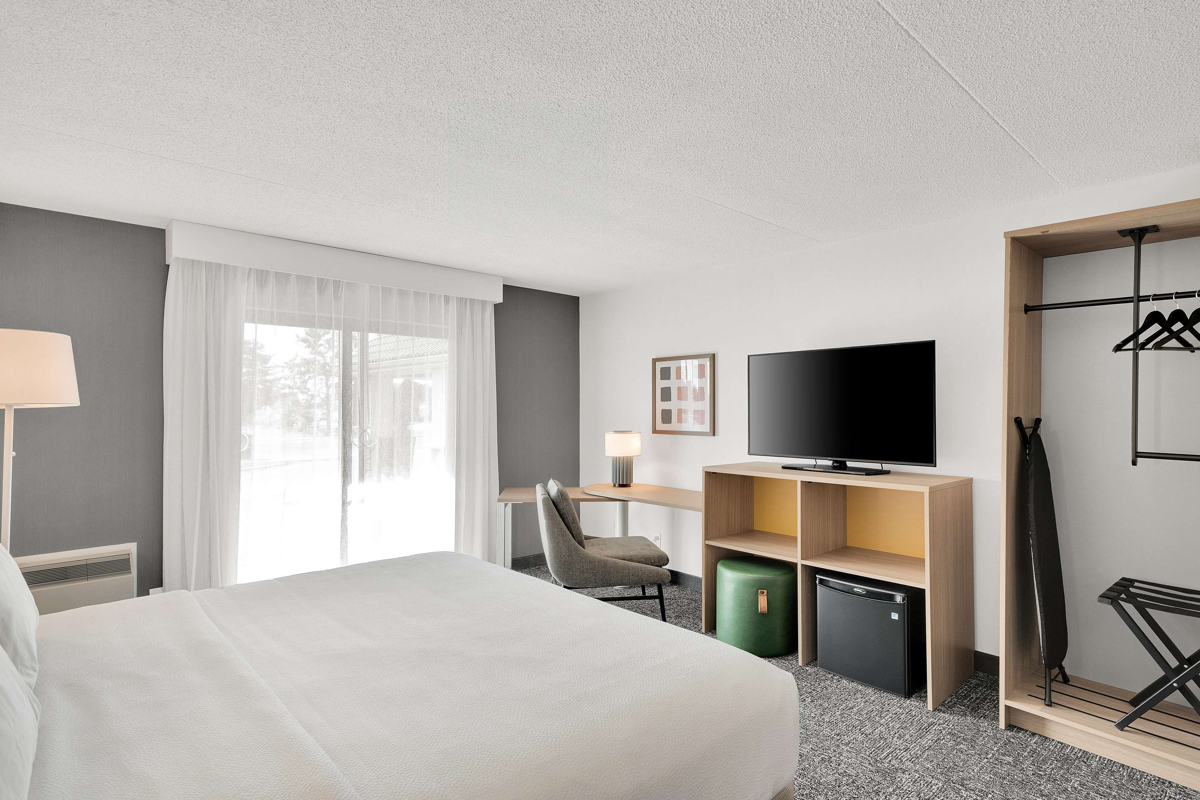 Habitación de hotel con cama, escritorio con silla, televisión, mini refrigerador y perchero.