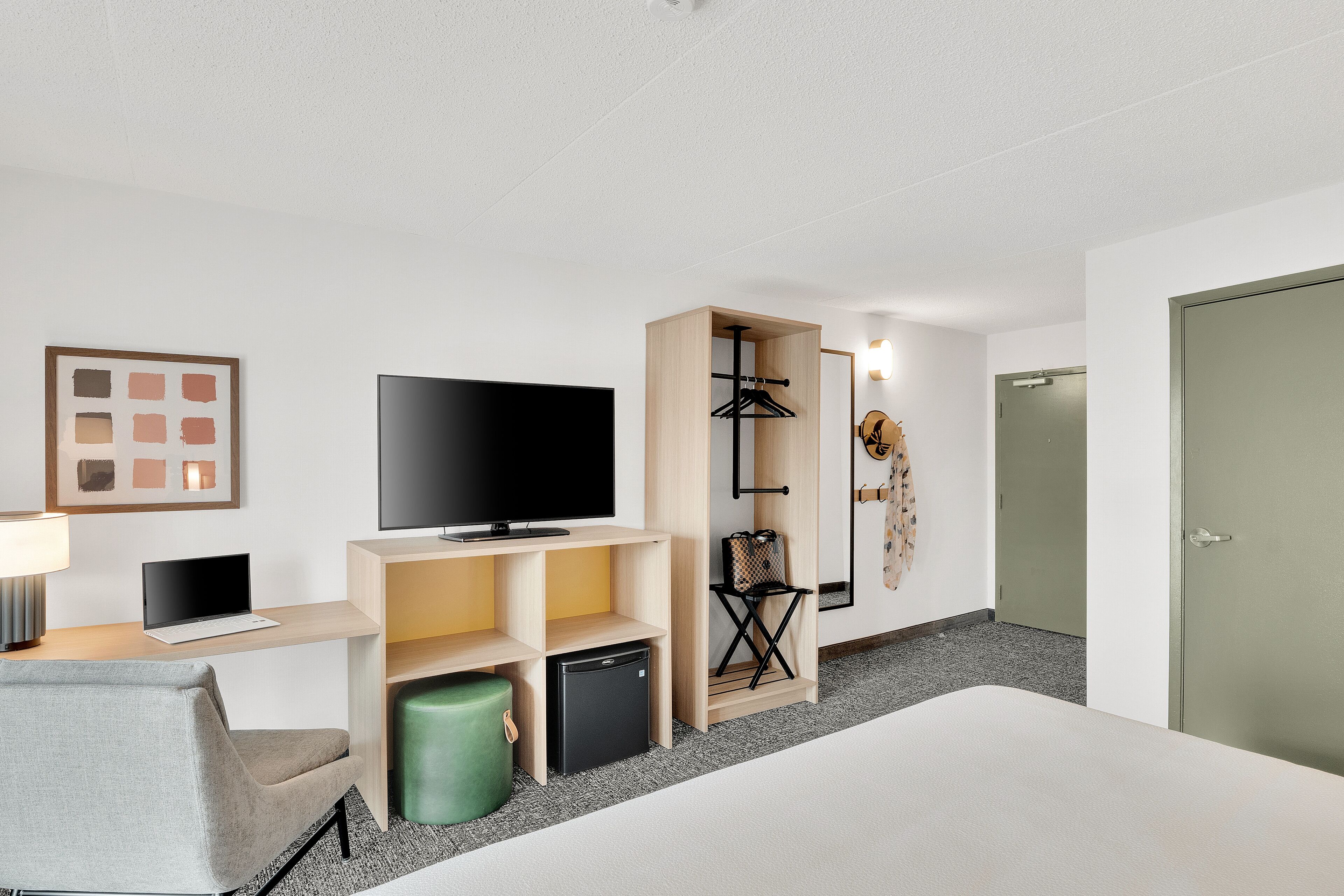 A hotel room with a bed, a TV, a desk with a laptop, a mini fridge, a green trash bin, a coat rack, and a door.