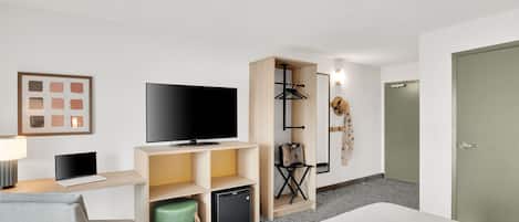A hotel room with a bed, a TV, a desk with a laptop, a mini fridge, a green trash bin, a coat rack, and a door.